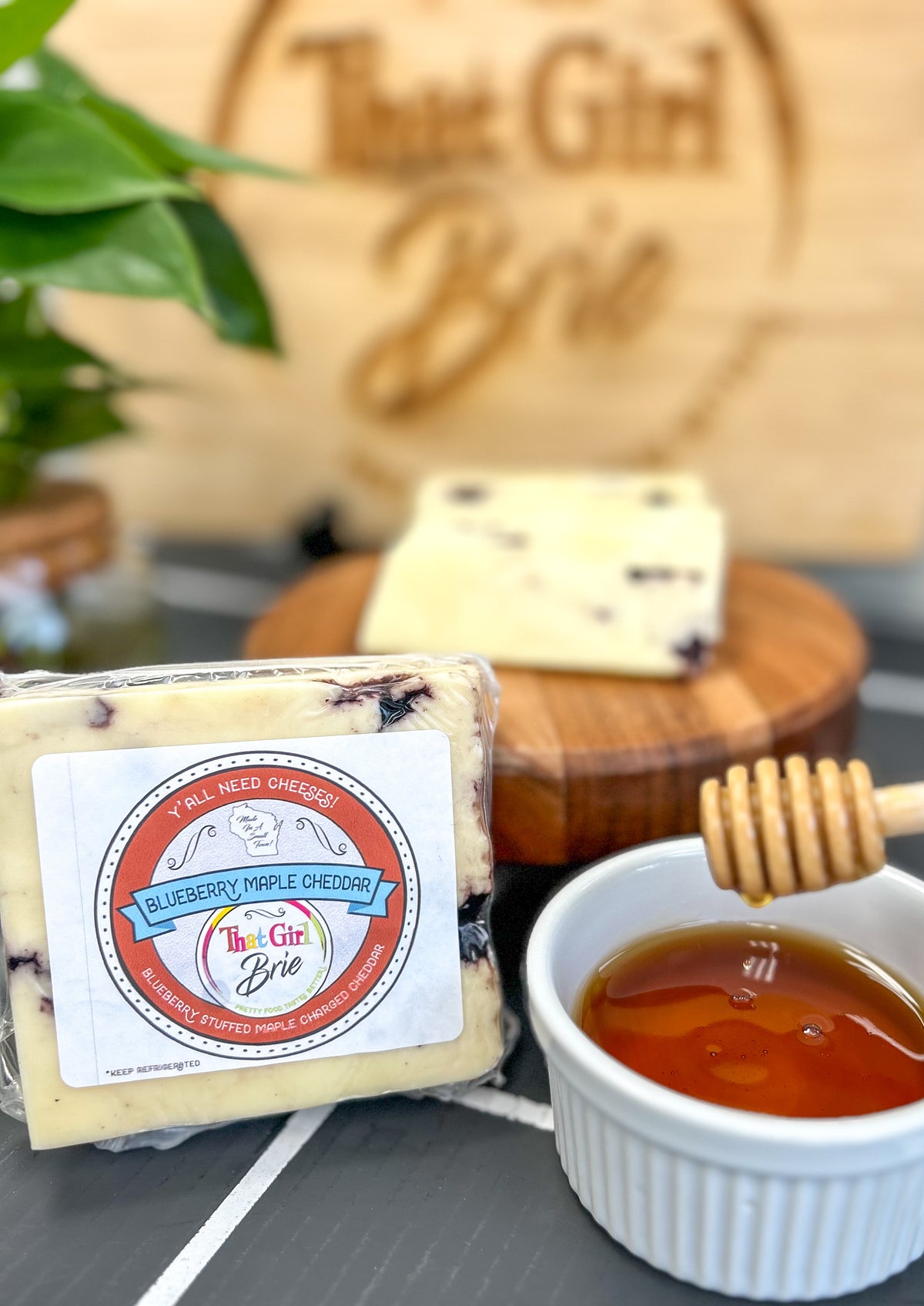 Blueberry Maple Cheddar blueberry cheddar cheese with a honey pot on a wood board in the That Girl Brie shop