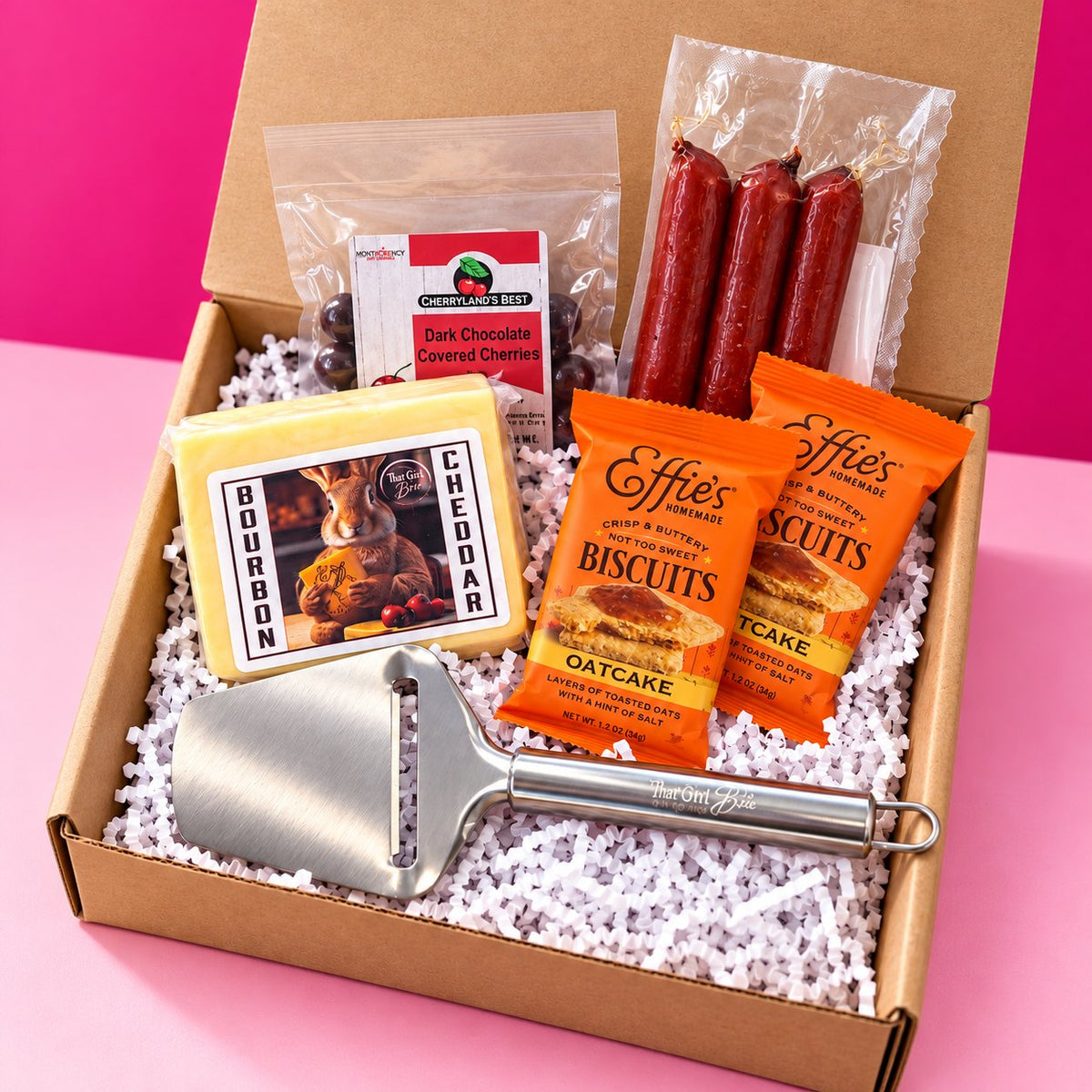 The Wisconsin Nightcap Wisconsin gift basket with a That Girl Brie cheese slicer on a hot pink background